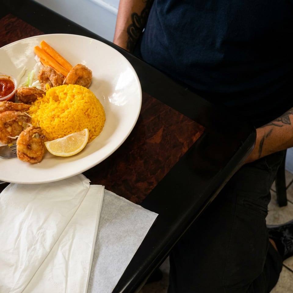 Plate of yellow rice, fried shrimp, vegetables, dipping sauce, and lemon beside utensils and a Coca-Cola can.